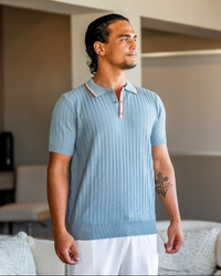 Man in a light blue ribbed knit polo with contrast striped collar and placket, front view with white trousers