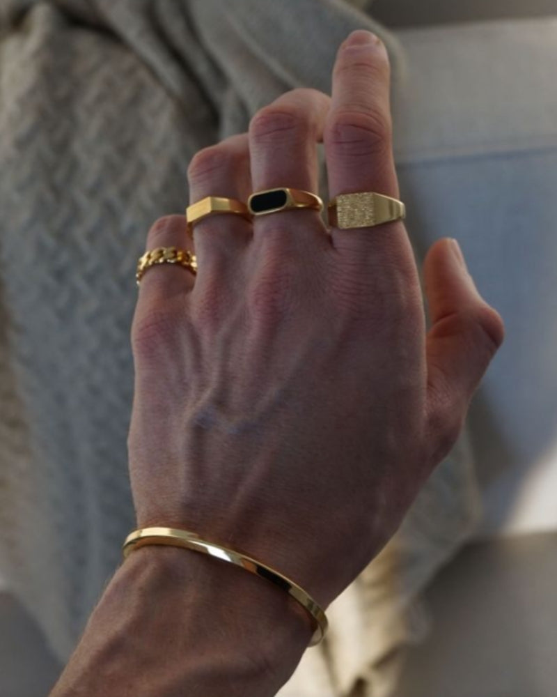 Hand wearing several gold-tone rings, including the black inlay band, and a gold cuff bracelet