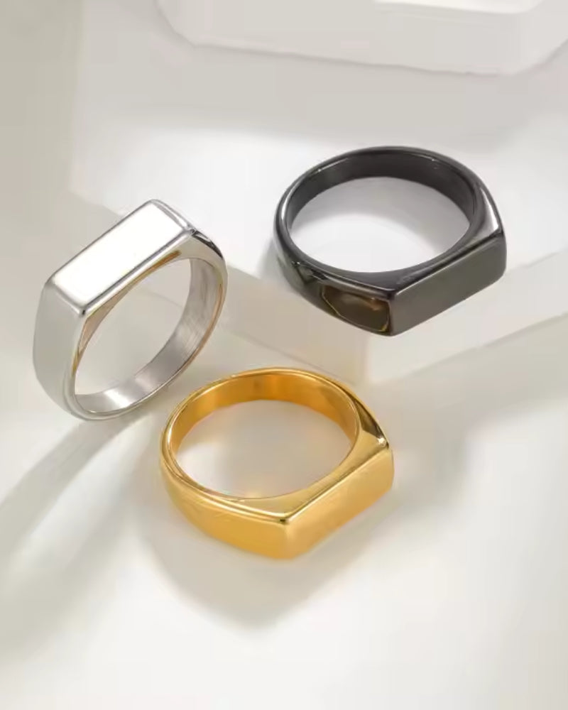 Rectangular signet rings in silver, black and gold finishes on a cream surface