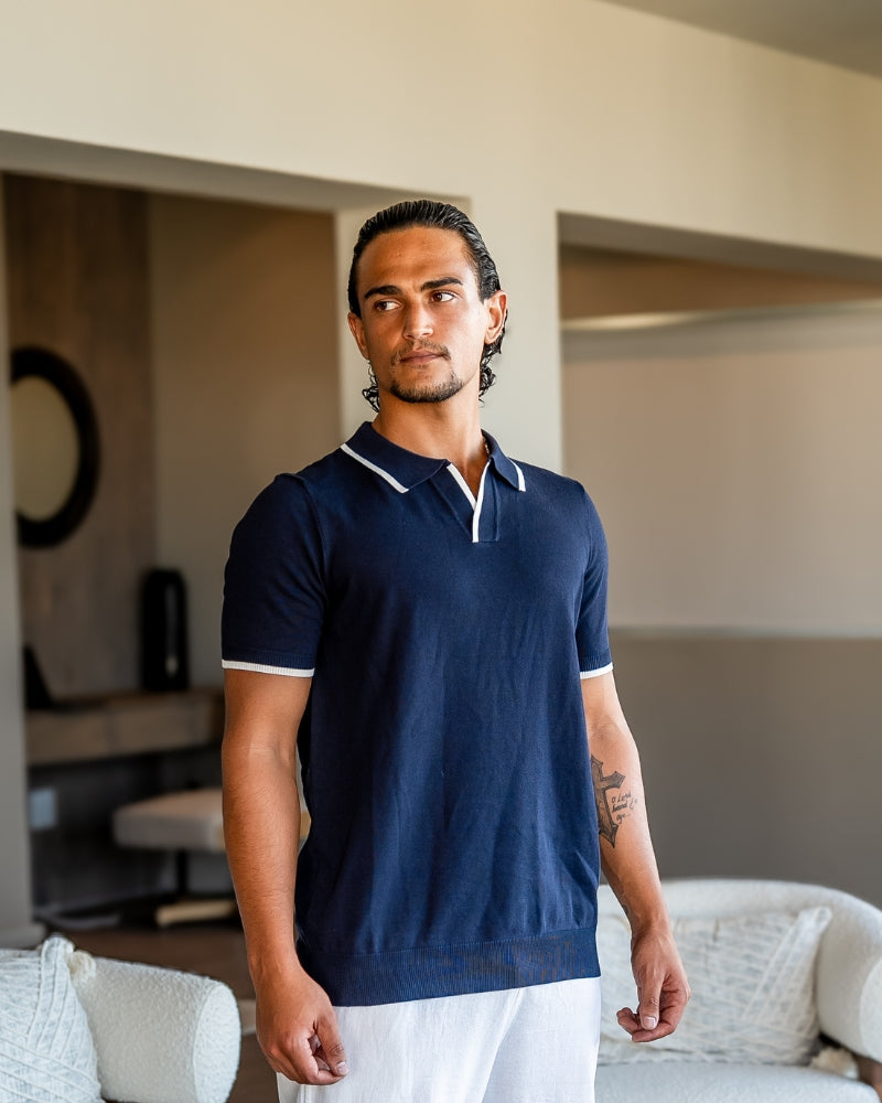 Man in a navy knit polo with white tipped collar and cuffs and white trousers, front view indoors