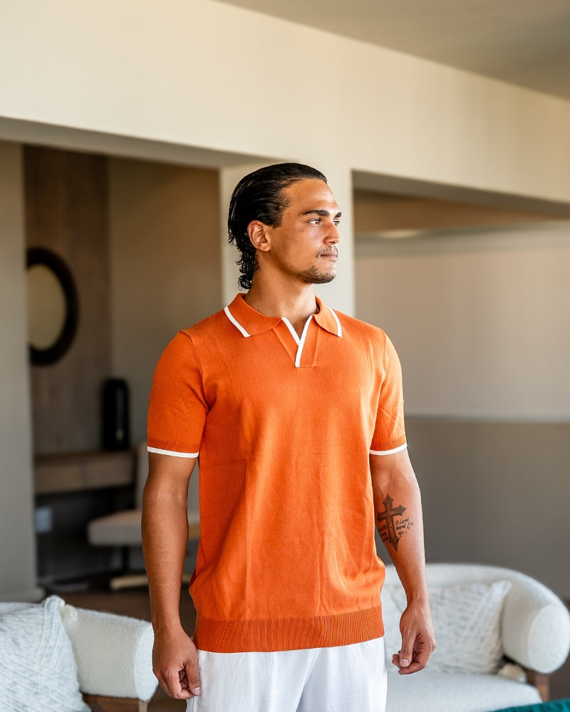 Man in a burnt orange knit polo with white tipping and white trousers, front view indoors