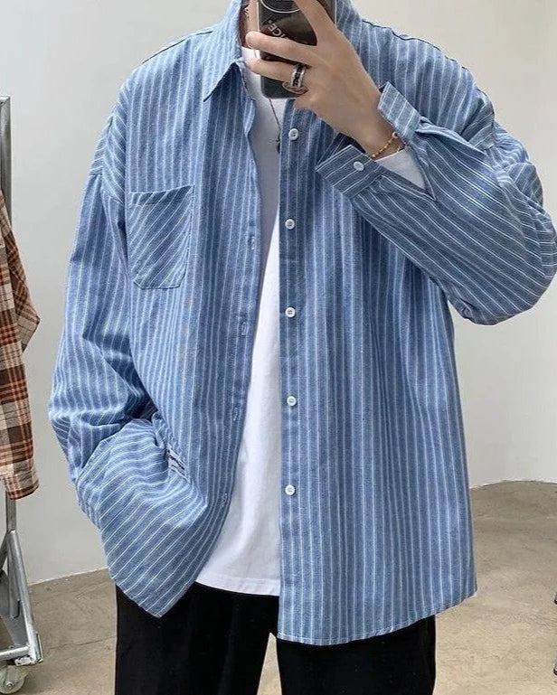Man in light blue striped oversized shirt over a white tee and black trousers, taking a mirror selfie