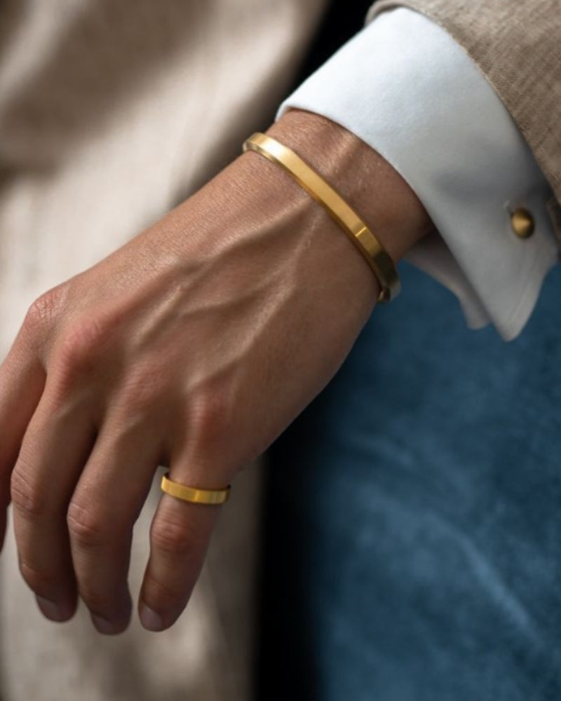 Man's wrist wearing a gold polished open cuff bracelet with a white shirt cuff and a gold ring