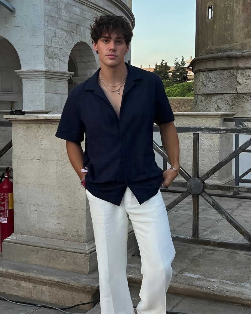 Man in navy short-sleeve camp shirt, white linen trousers and layered gold chains, standing by a stone pillar