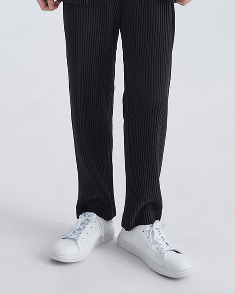 Close-up of black fine-pleated trousers from the knee down with white trainers, front view