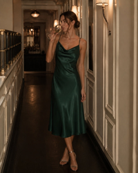 Woman in emerald green satin cowl-neck slip dress holding a glass in a corridor