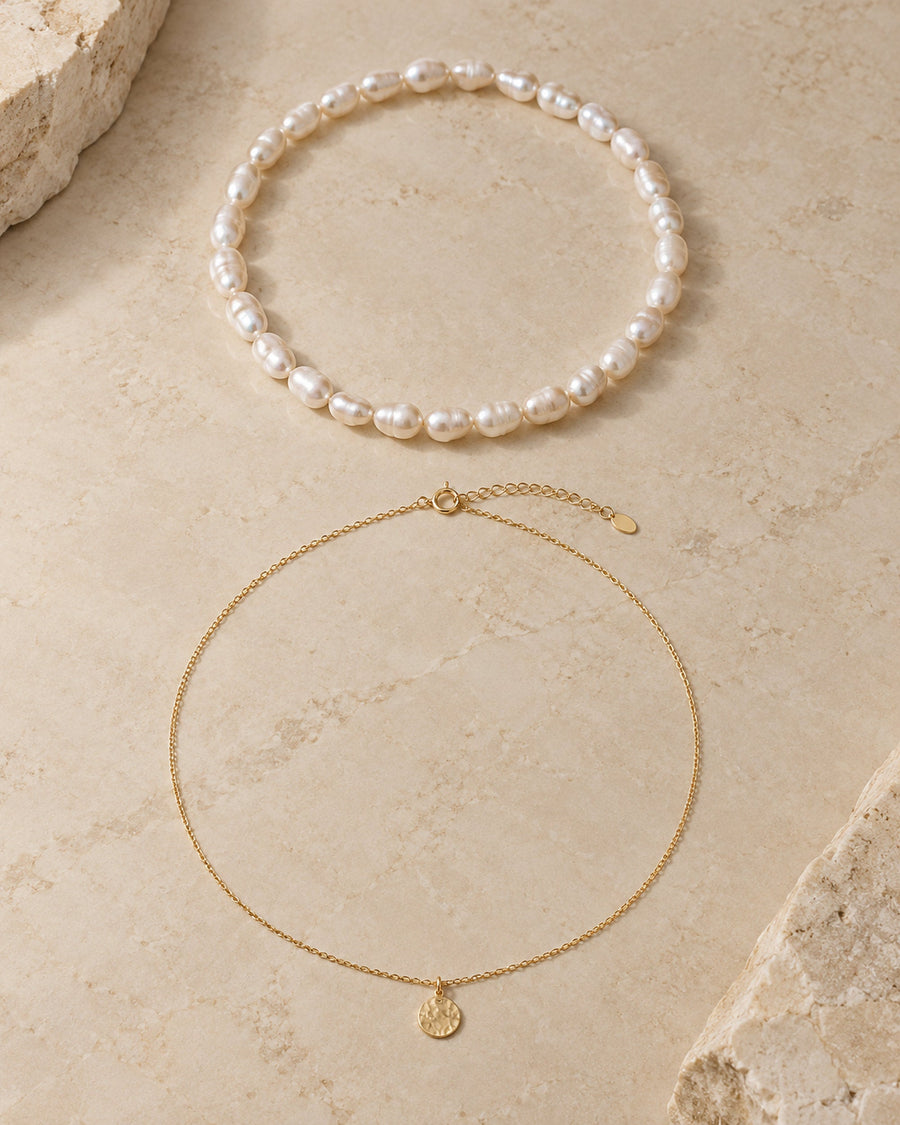 Pearl choker and gold-tone coin pendant necklace laid flat on pale stone