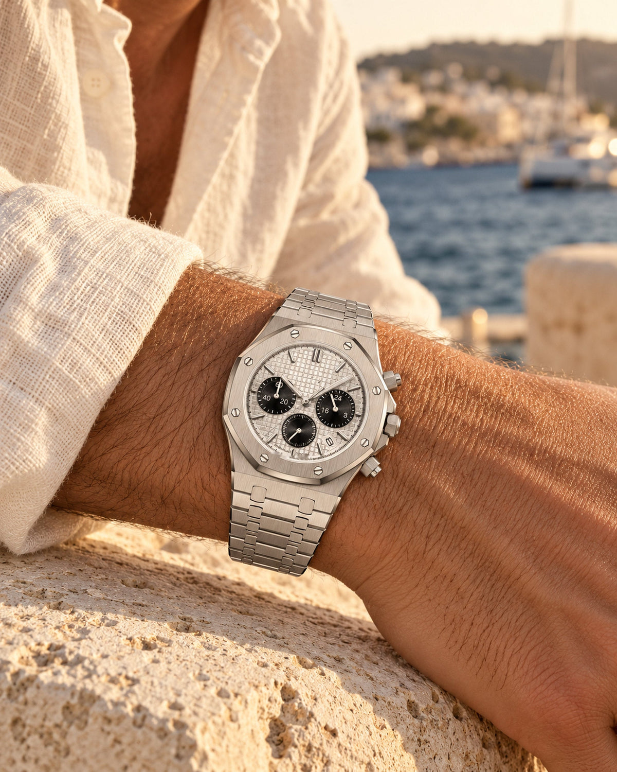 Man's wrist wearing a silver chronograph watch with octagonal bezel beside a sunlit harbour
