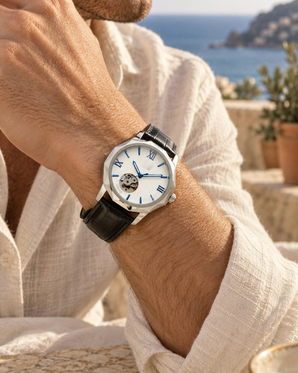 Man's wrist wearing a skeleton dial watch with black leather strap and white linen shirt by the sea