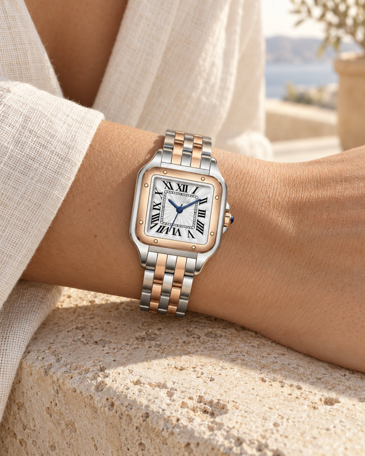 Two-tone square watch with rose-gold bezel on a wrist, linen sleeve and sea view behind