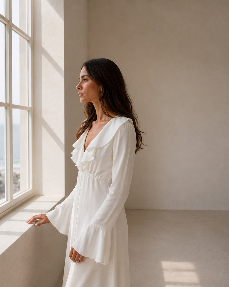Side profile of woman in ivory ruffle-collar dress with bell sleeves, looking out of a window