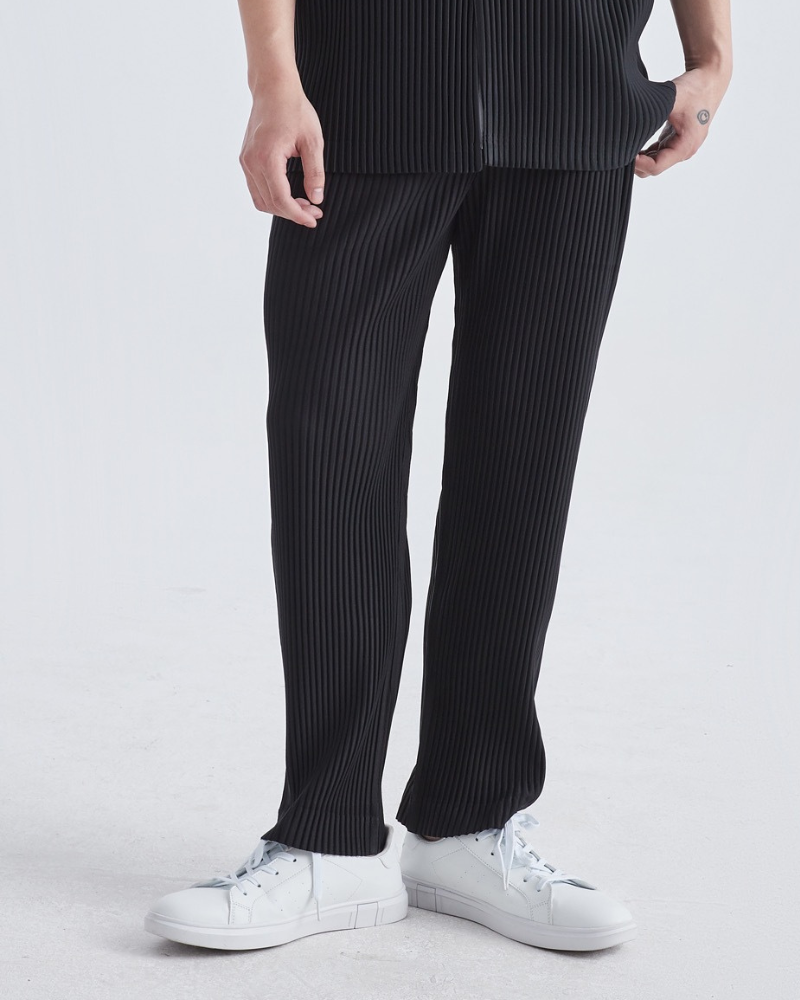 Man in black fine-pleated straight trousers and white trainers, cropped front view on grey background