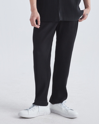 Man in black fine-pleated straight trousers and white trainers, cropped front view on grey background