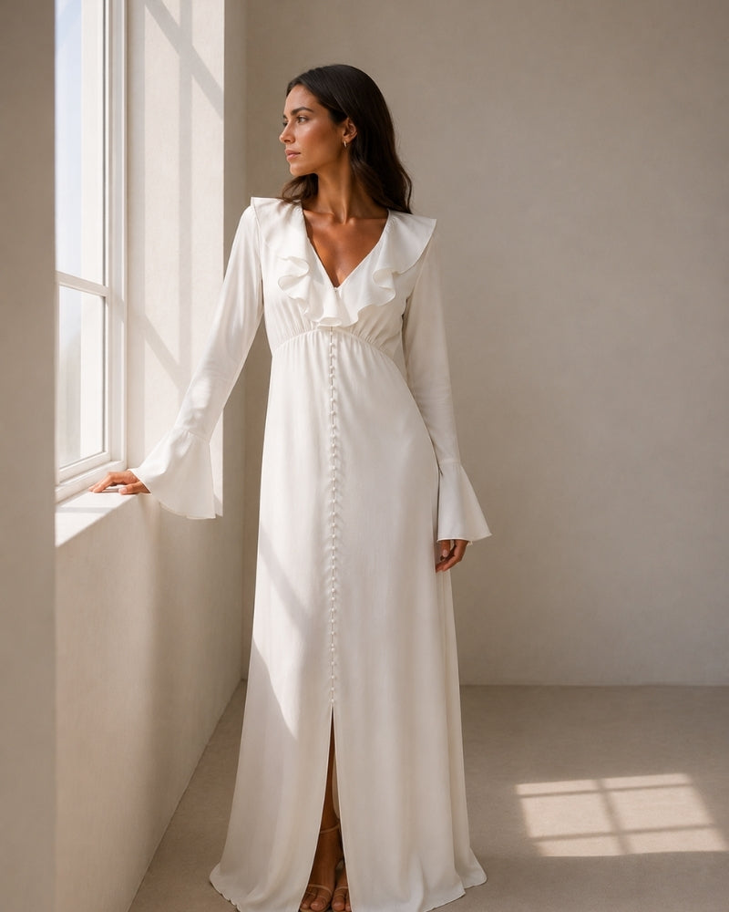 Woman in ivory ruffle-collar maxi dress with pearl button front and bell sleeves, standing by a window
