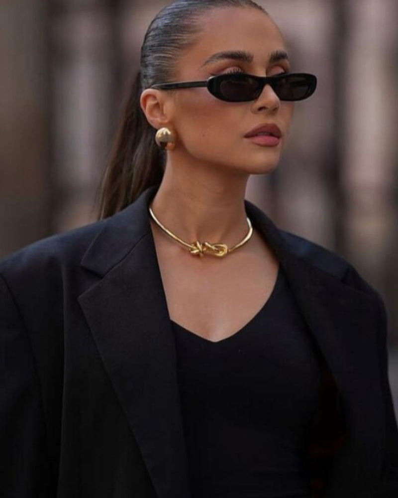 Woman wearing black slim oval sunglasses with a black blazer and gold collar necklace