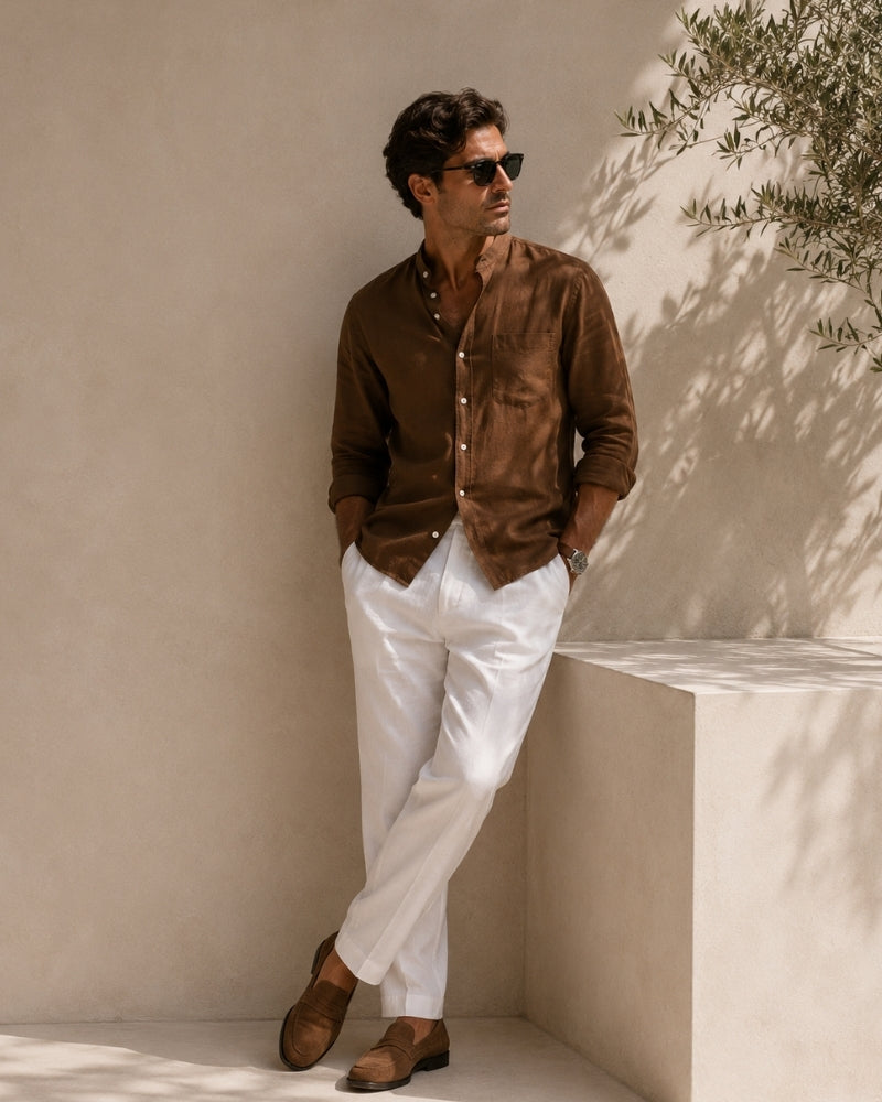 Man in sunglasses leaning on a pale wall in brown mandarin collar shirt and white trousers