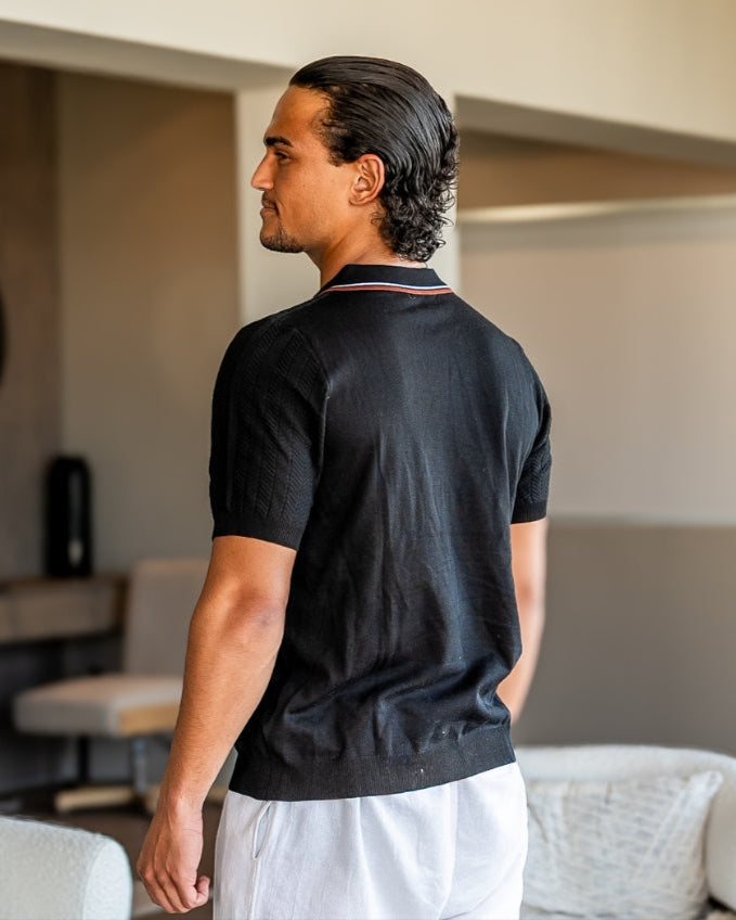 Vue de dos d'un polo en maille côtelée noir à col rayé, porté avec un pantalon blanc