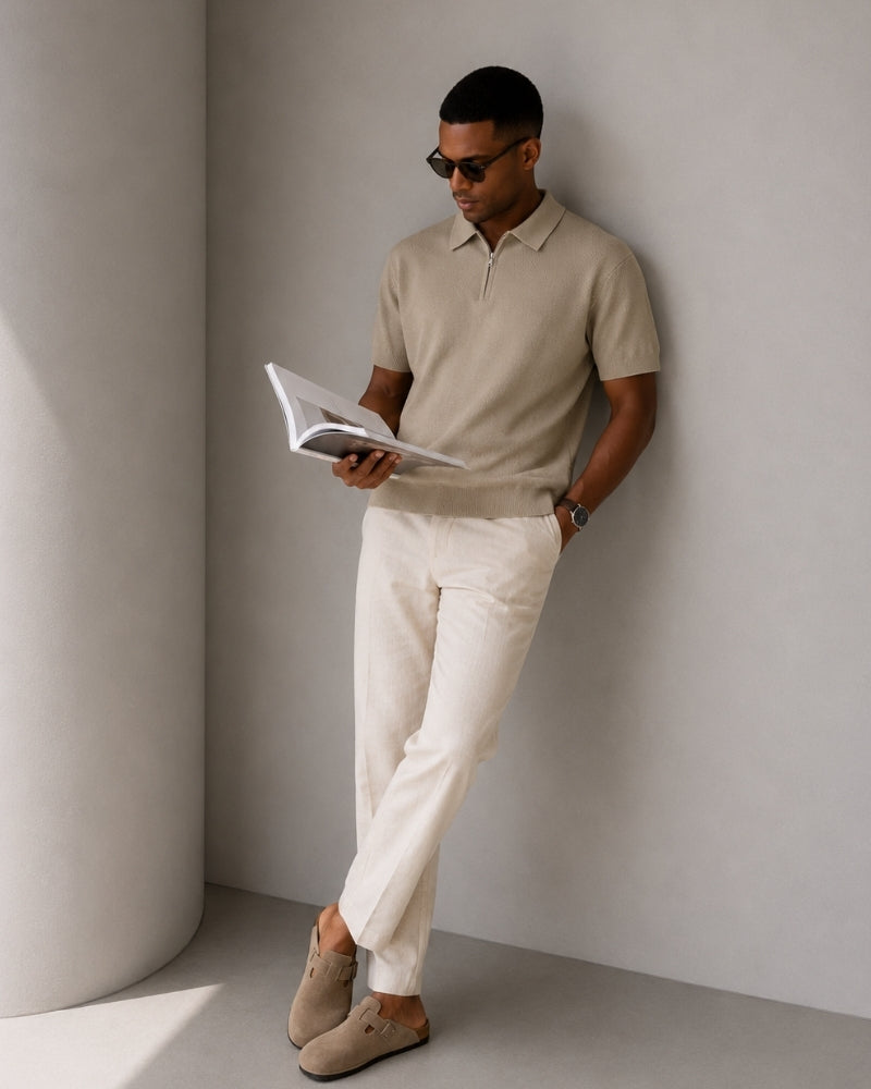 Man in sunglasses leaning on a wall reading, wearing taupe zip polo and cream trousers