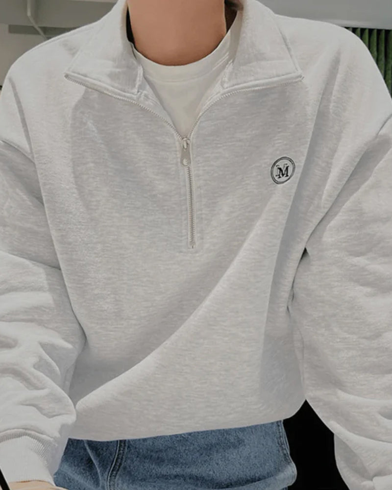 Light grey marl half-zip sweatshirt with round M logo worn over a white tee and jeans, close-up