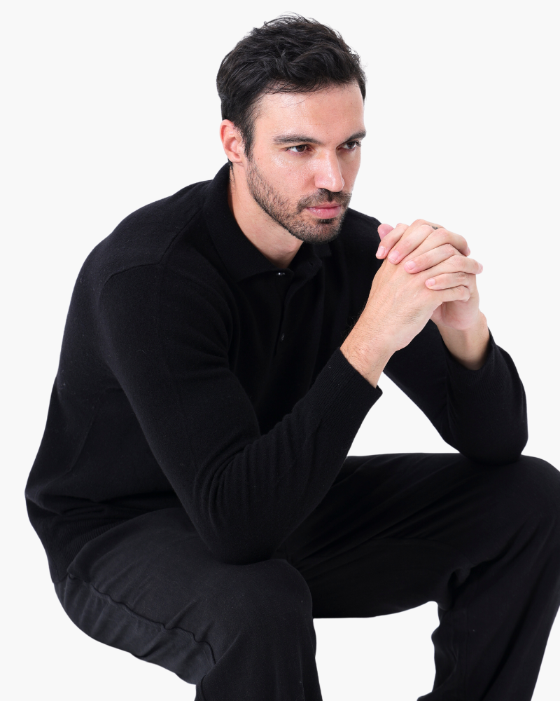 Man in black long-sleeve polo jumper and black trousers, seated with hands clasped
