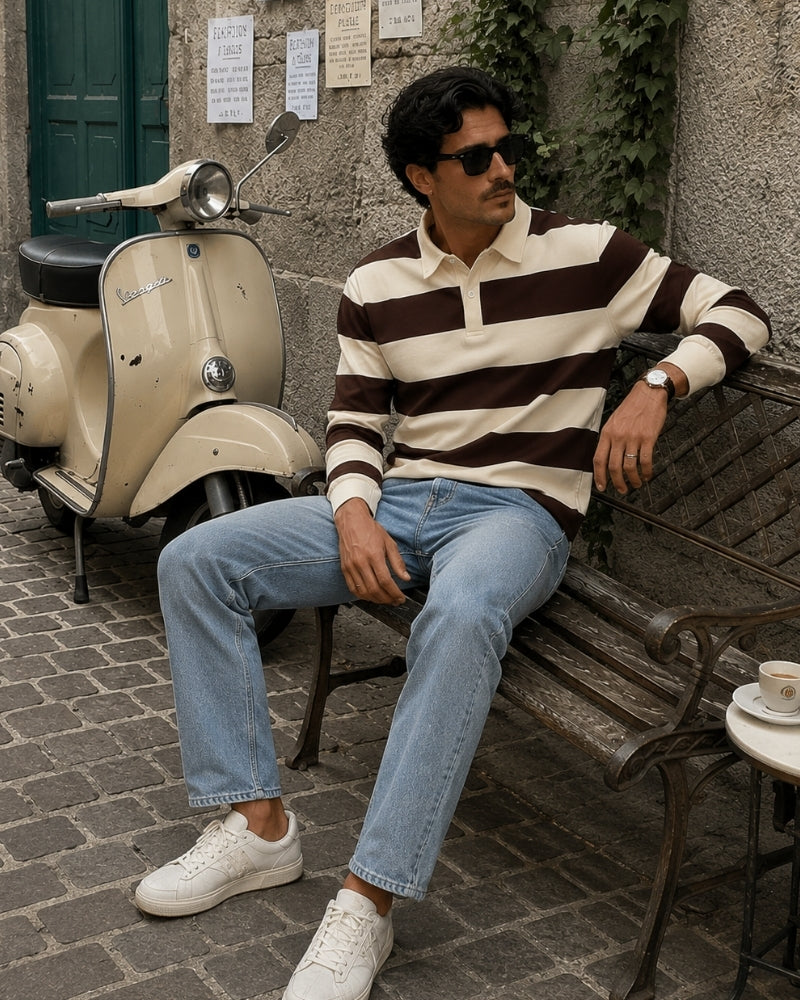 Man in brown and cream striped rugby polo with blue jeans, seated on a bench beside a scooter