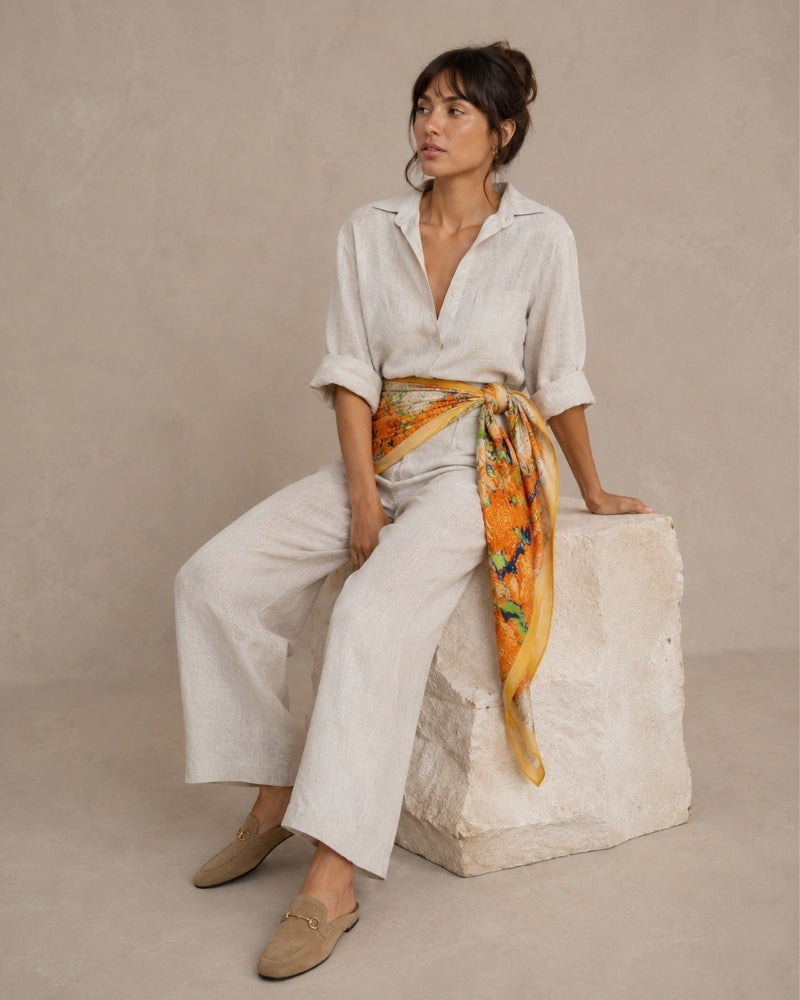 Woman in cream linen outfit seated on stone, orange floral scarf tied at the waist