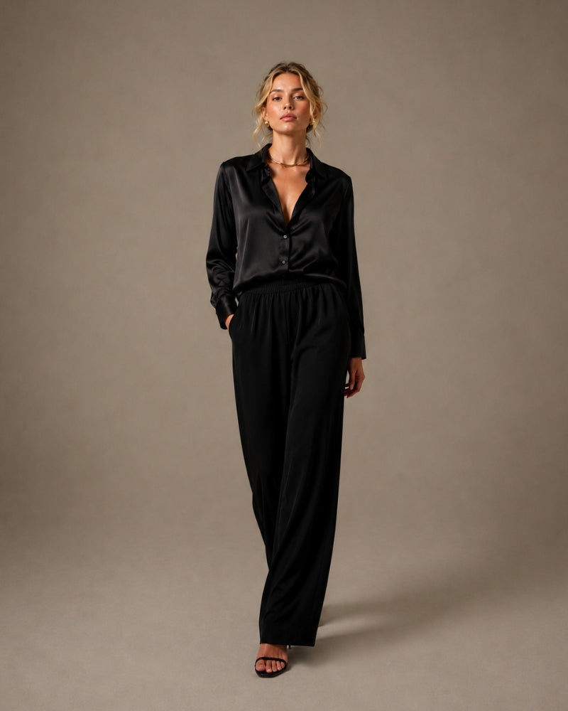 Femme en chemise en satin noire et pantalon large noir, une main dans la poche, fond de studio gris