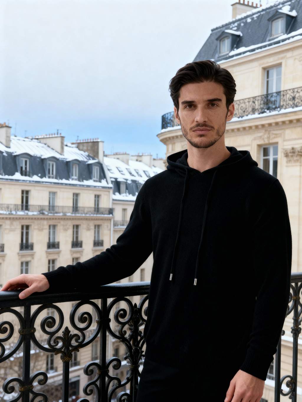 Man in a black cashmere hoodie on a snowy Paris balcony, drawstring hood and relaxed fit