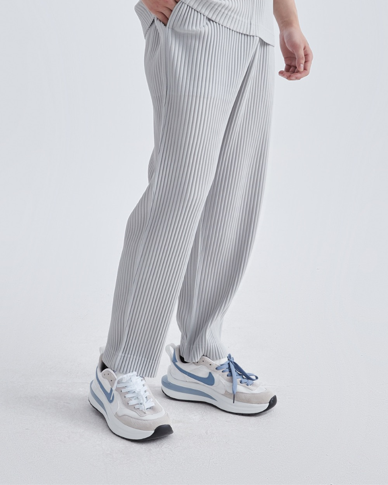 Side view of man in light grey fine-pleated trousers with white-and-blue trainers, hand in pocket