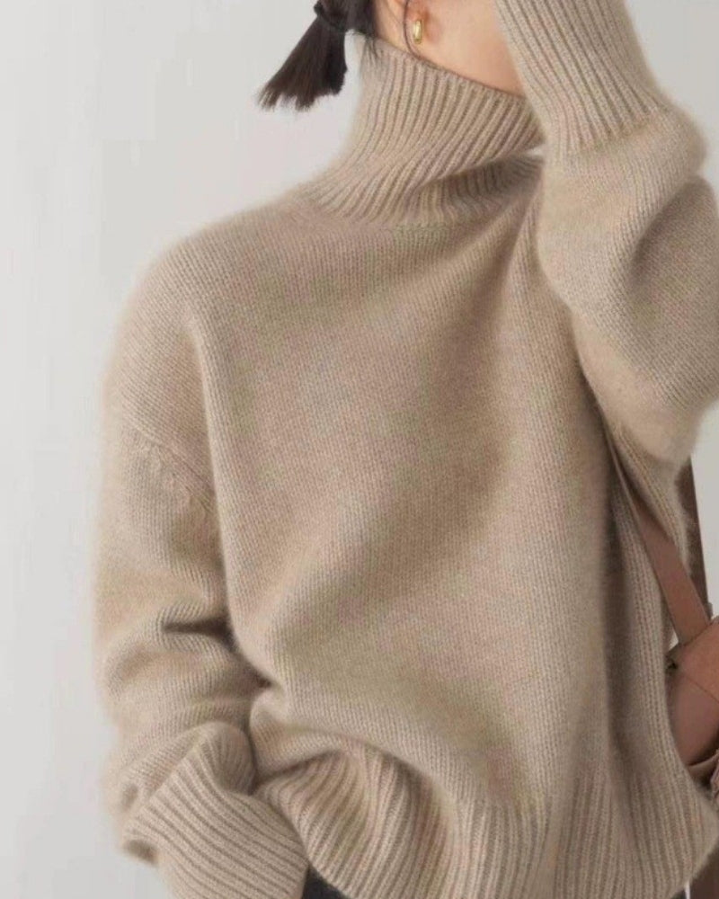 Woman wearing oversized beige turtleneck knit, hand raised to collar, brown bag strap