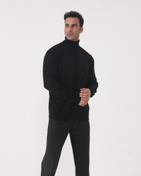 Hombre con jersey de cuello alto negro y pantalón gris, manos entrelazadas, de pie