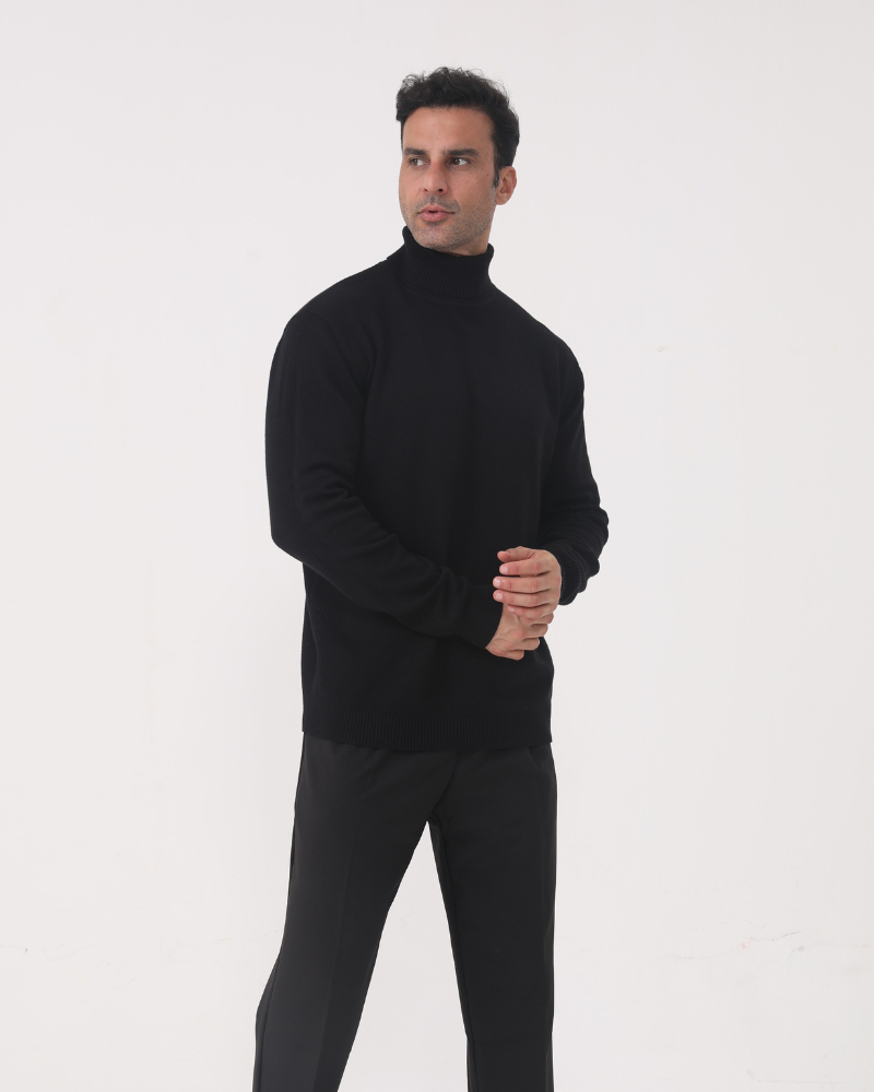 Man wearing black turtleneck jumper with grey trousers, hands clasped, standing