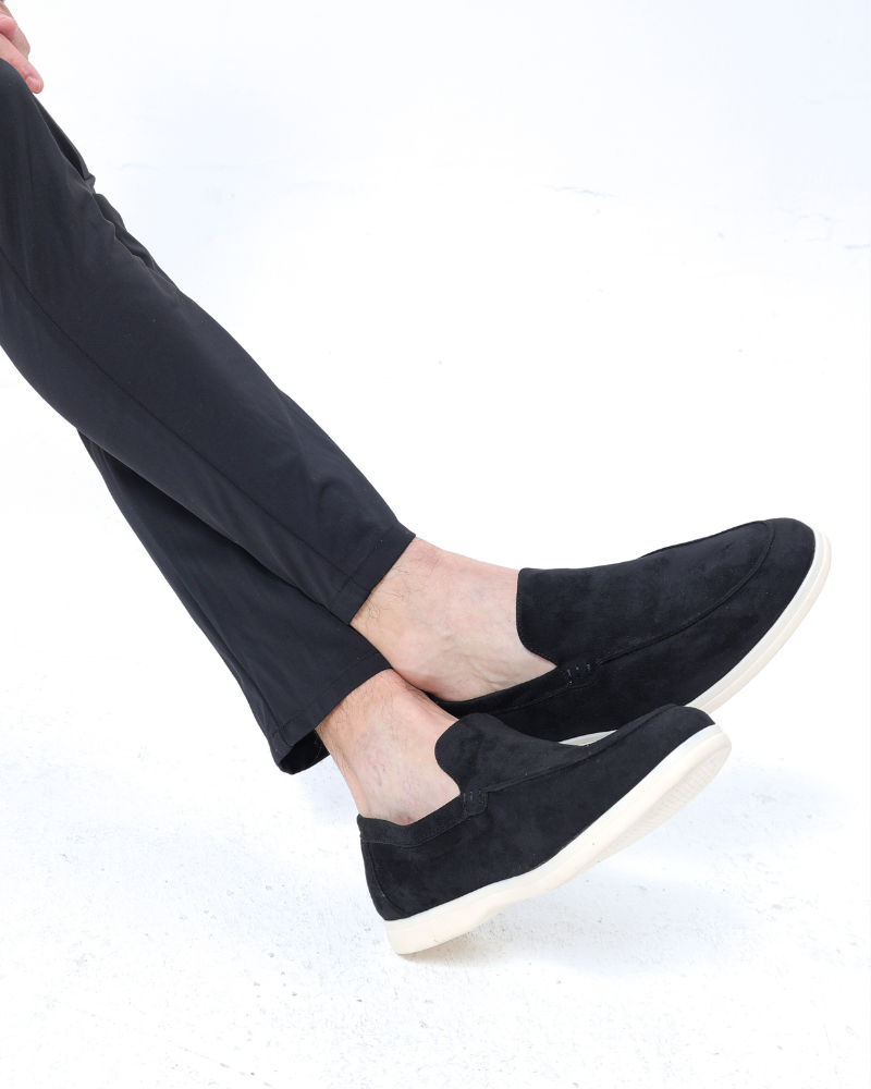 Legs crossed in black trousers and black suede loafers with cream soles, side view on white