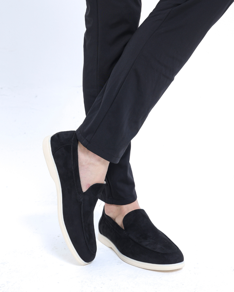 Man in black trousers and black suede loafers with cream soles, legs crossed, front view on white