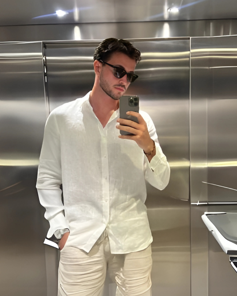 Man in white linen shirt and black sunglasses taking a mirror selfie in a steel lift