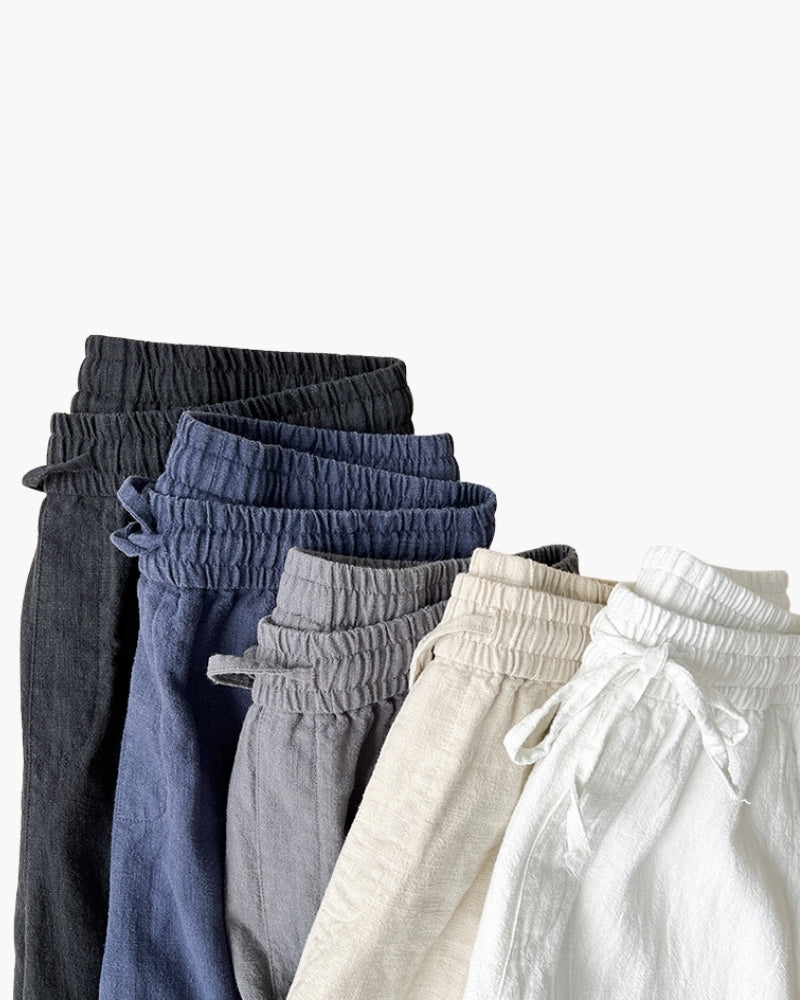 Pila de pantalones de lino con cordón doblados en negro, azul marino, gris, avena y blanco con las cinturas elásticas a la vista