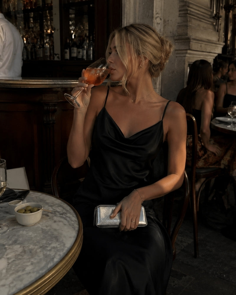 Femme en robe nuisette en satin noir à col bénitier tenant un verre à une table de café
