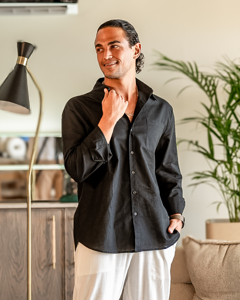 Man in an open-collar black linen shirt and white trousers, smiling with a hand at the collar, indoors