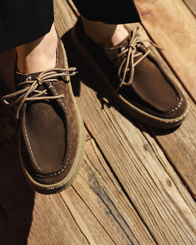 Close-up of dark brown suede moc-toe lace-ups with crepe soles worn on wooden decking, top-down view