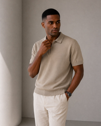 Man wearing taupe knit zip polo with cream trousers, hand at collar against grey wall