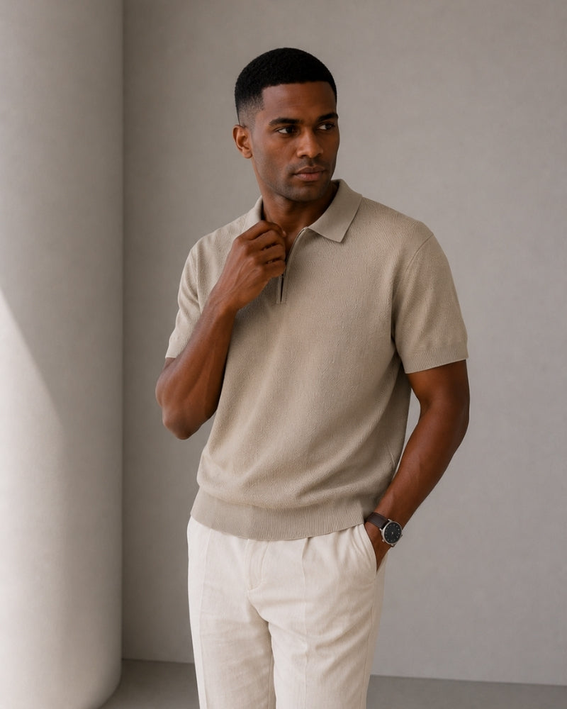 Man wearing taupe knit zip polo with cream trousers, hand at collar against grey wall