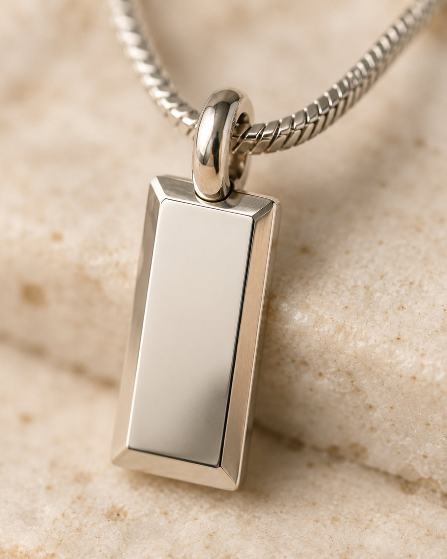 Close-up of a silver rectangular tag pendant with bevelled edges resting on sandy stone