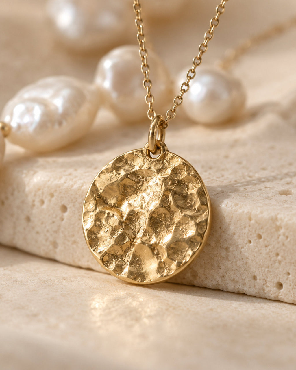 Primer plano de un colgante de moneda martillado dorado en cadena junto a perlas de agua dulce sobre piedra
