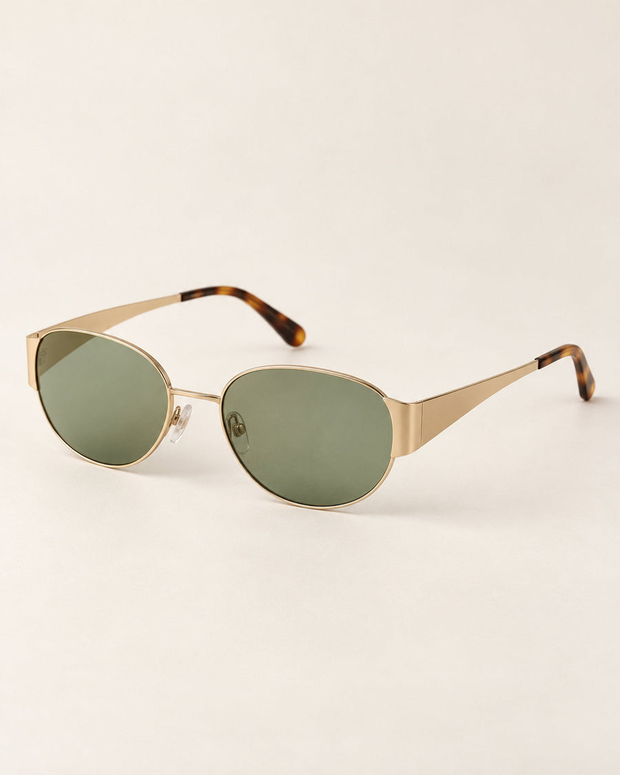 Gold-tone oval sunglasses with green lenses and tortoiseshell tips, angled view on a beige surface