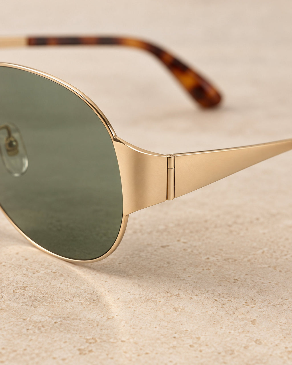 Close-up of green sunglass lens, gold-tone hinge and tortoiseshell temple tip on sand