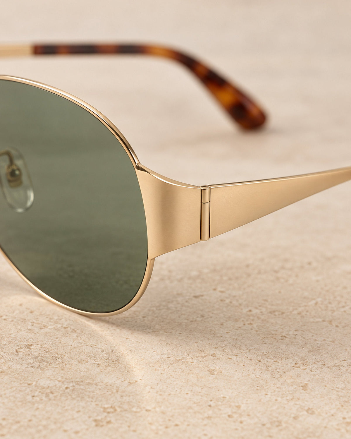 Close-up of green sunglass lens, gold-tone hinge and tortoiseshell temple tip on sand