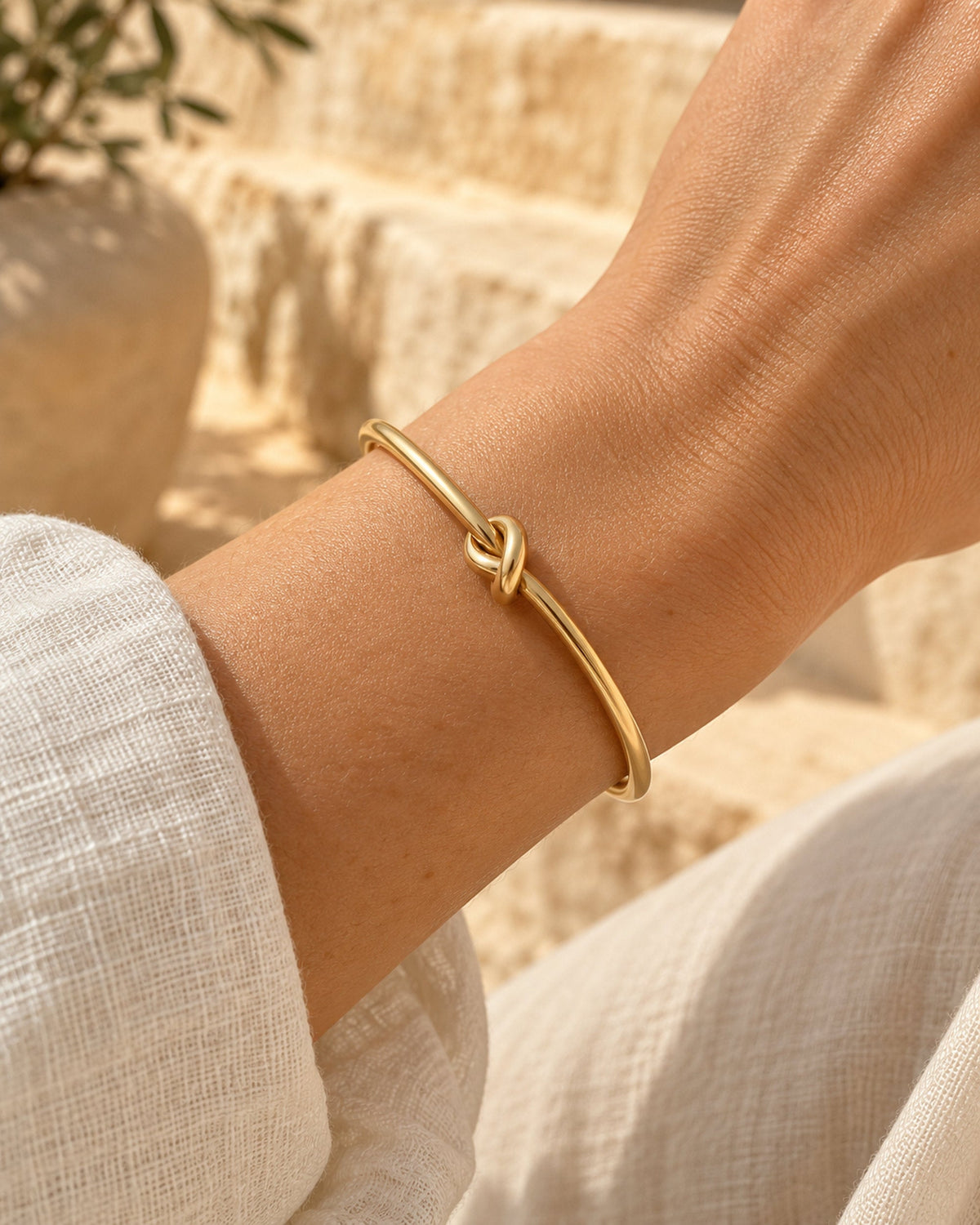Woman wearing gold-tone knot cuff bracelet on wrist, cream linen sleeve, stone steps behind