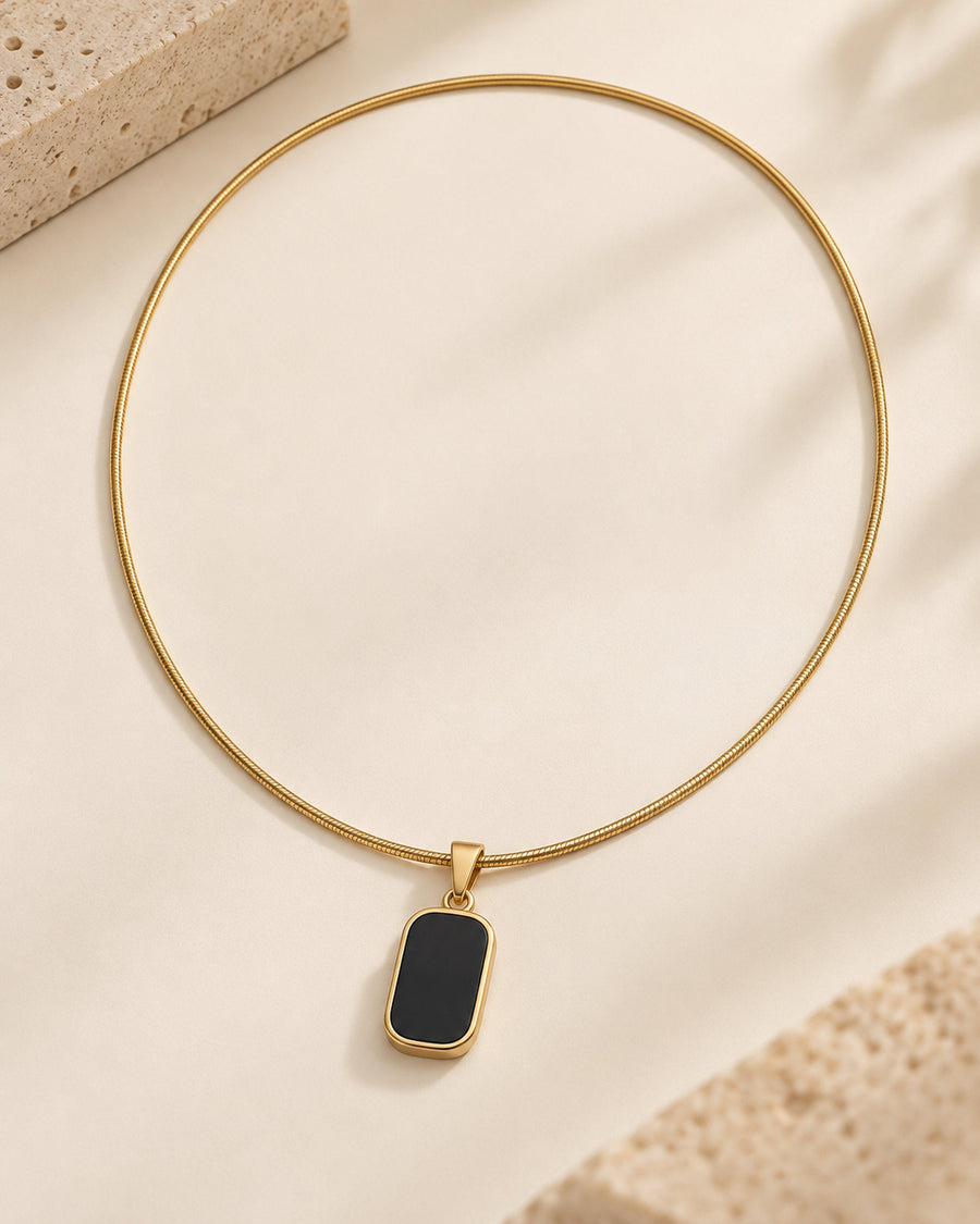 Gold-tone snake chain necklace with black rectangular pendant, flat-lay on cream surface beside stone
