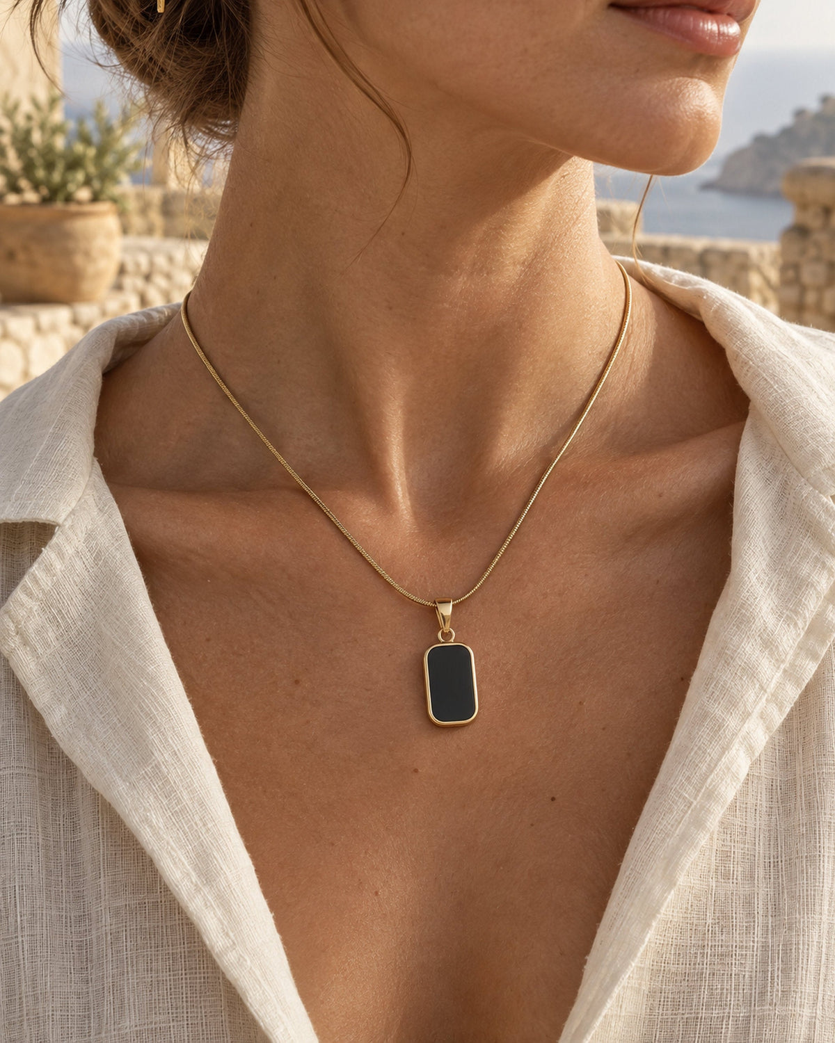 Woman wearing gold-tone snake chain necklace with black rectangular pendant over a linen shirt