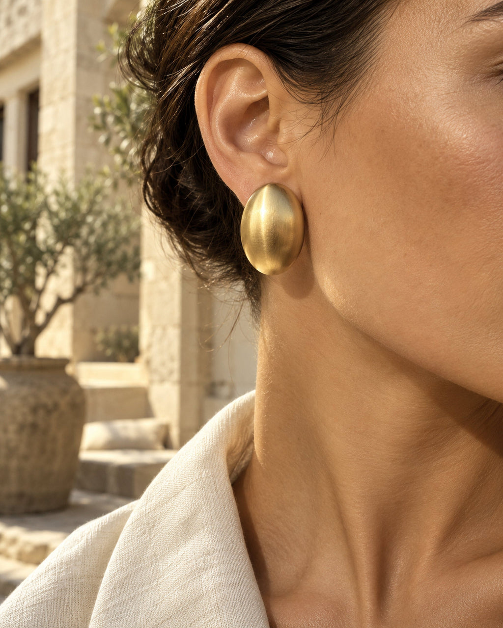 Femme portant une boucle d'oreille ovale dorée brossée, olivier et col en lin derrière, gros plan